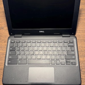 May include: Black Dell laptop with an open screen, revealing a keyboard and touchpad. The laptop is on a wooden surface. The Dell logo is visible above the keyboard.