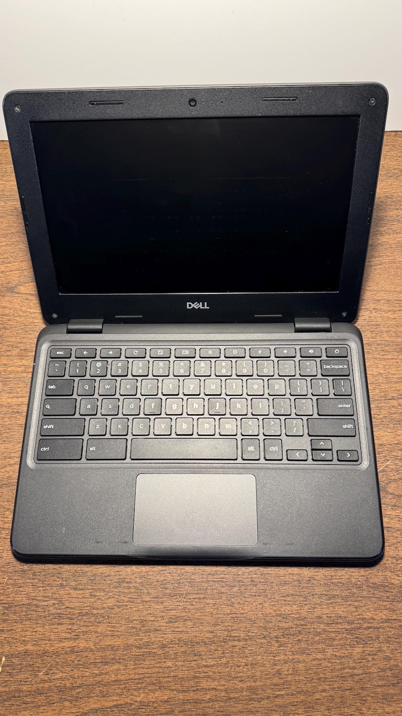 May include: Black Dell laptop with an open screen, revealing a keyboard and touchpad. The laptop is on a wooden surface. The Dell logo is visible above the keyboard.