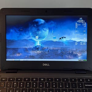 Könnte beinhalten: Ein schwarzes Dell Chromebook-Laptop mit Tastatur und einem Bildschirm, der eine blau-weiße Landschaft mit einer großen Kugel zeigt. Der Bildschirm zeigt auch Symbole und Systeminformationen an. Der Laptop ist geöffnet.