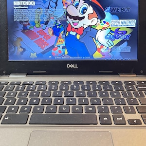 May include: A Dell laptop displaying a Super Nintendo Entertainment System (SNES) game interface. The screen shows a colorful Mario illustration and game titles. The keyboard is visible below.