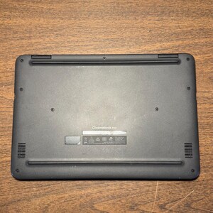 May include: A black Chromebook 3100 laptop, viewed from the bottom, rests on a wooden surface. The laptop has a smooth, matte finish with vents and small circular details. The text "Chromebook 3100" is visible.