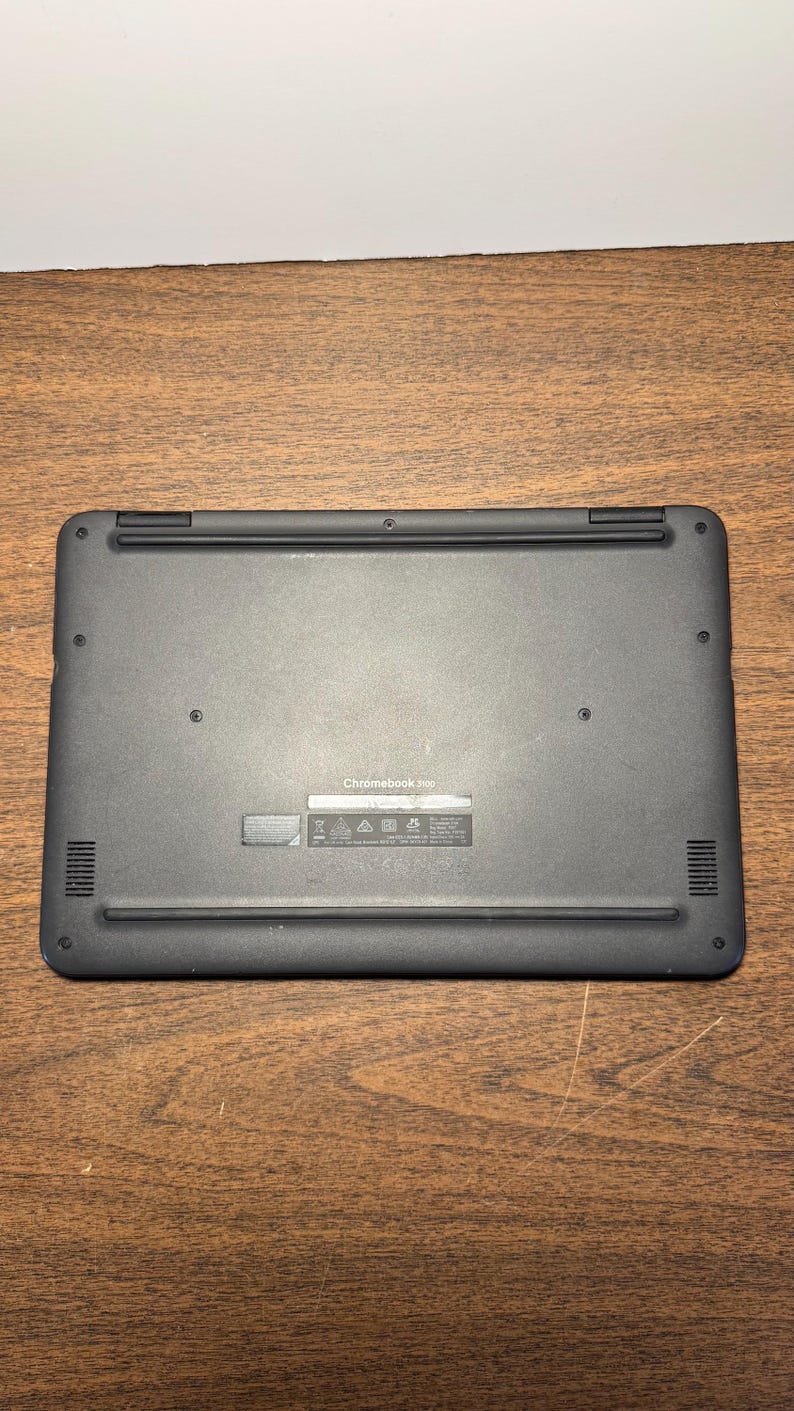 May include: A black Chromebook 3100 laptop, viewed from the bottom, rests on a wooden surface. The laptop has a smooth, matte finish with vents and small circular details. The text "Chromebook 3100" is visible.