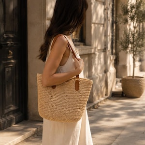 May include: A woven straw tote bag with brown leather handles is carried by a person wearing a cream-colored dress. The bag has a rectangular shape and a textured surface, perfect for a day out.