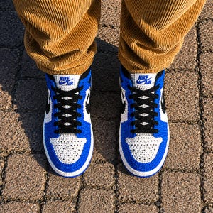 May include: A pair of crochet sneakers featuring a white and royal blue color scheme with black laces and a black swoosh logo. The tongue features a white label with the blue text Nike Air. The sneakers are worn with tan corduroy trousers.