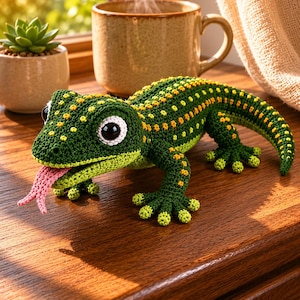 Puede incluir: Un juguete de gecko tejido a ganchillo con cuerpo verde oscuro y manchas amarillas, grandes ojos negros y blancos, y una lengua rosa que sobresale. El lagarto tiene cuatro patas con dedos definidos y una cola larga y curva de hilo.