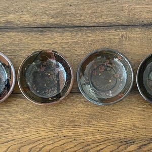 May include: A set of six circular ceramic bowls arranged in a horizontal row. Each bowl features a unique glazed finish with varying shades of dark brown, black, and grey, accented by subtle speckled patterns and smooth, reflective surfaces.