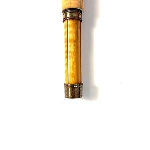 Peut inclure: Gros plan sur le manche d'une canne &agrave; p&ecirc;che &agrave; la mouche. La poign&eacute;e est en li&egrave;ge et en bois clair, avec des accents m&eacute;talliques de couleur laiton. La canne est pos&eacute;e sur un fond blanc.