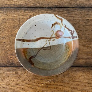 May include: A round ceramic bowl featuring a two-tone glaze design. The upper portion is a speckled off-white, while the lower section is a deep brown. A dark brown, hand-painted abstract line pattern crosses the centre, accented by a small reddish-brown circular detail.