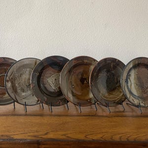 May include: A set of six round ceramic plates displayed on black metal stands. Each plate features a unique, mottled glaze in shades of brown, black, and tan, with visible swirling patterns, textured surfaces, and irregular dark markings across the centres.