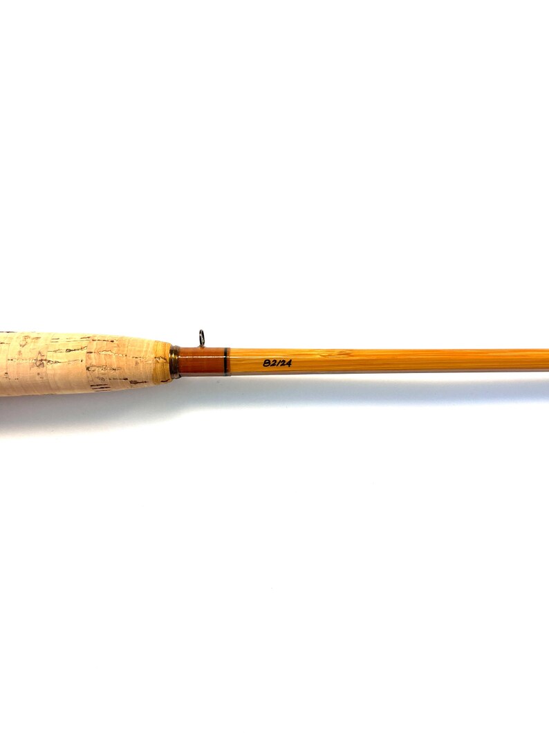 Peut inclure: Canne &agrave; p&ecirc;che &agrave; la mouche vintage avec une poign&eacute;e en li&egrave;ge et un f&ucirc;t en bambou brun clair. La canne a un accent marron fonc&eacute; pr&egrave;s de la poign&eacute;e et le texte "B2/24" imprim&eacute; sur le f&ucirc;t. Sur fond blanc.