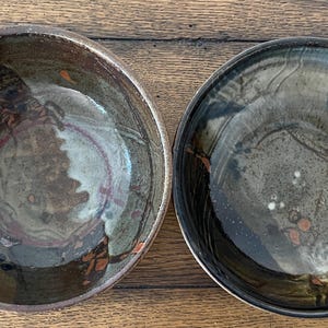 May include: Two round ceramic bowls with a glossy finish, featuring a dark brown and black glaze with subtle grey and cream-coloured marbling. The interior surfaces display abstract patterns with small orange and dark blue accents throughout.