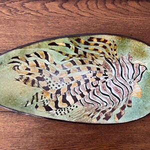 May include: An oval-shaped ceramic platter featuring a detailed hand-painted illustration of a lionfish. The fish displays prominent striped fins and spines in shades of brown, black, and cream, set against a mottled sage green background.