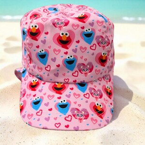May include: A pink baseball cap featuring an all-over print of Sesame Street characters Elmo, Cookie Monster, and Abby Cadabby, each framed within heart shapes, interspersed with small red, purple, and pink hearts across the entire fabric surface.