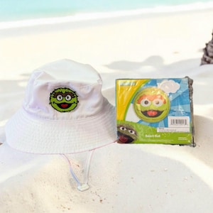 May include: A white bucket hat with a green and black cartoon character patch, and a beach ball in packaging. The beach ball features the same character. The items are on a sandy beach.