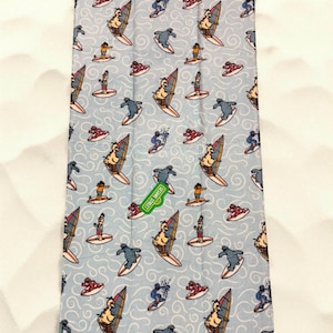 May include: A light blue rectangular towel featuring a repeating pattern of Sesame Street characters windsurfing and surfing on waves. A green rectangular tag with the text Sesame Street is attached to the center of the fabric surface.