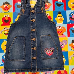 May include: A denim overall dress with a button-up front and two front pockets. The dress features a colorful embroidered design of a Sesame Street character on one pocket. The dress has adjustable straps and is suitable for children.