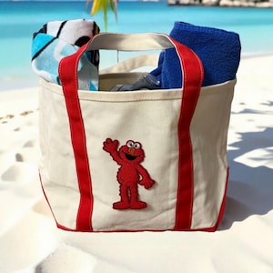 May include: A canvas tote bag with red accents and a red Elmo patch. The bag is filled with blue and patterned towels, perfect for a day at the beach. The bag is sitting on white sand with a beach and ocean in the background.