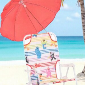 May include: A red beach umbrella provides shade over a folding beach chair. The chair features a colorful pattern with cartoon characters. The scene is set on a sandy beach with a palm tree and ocean in the background.