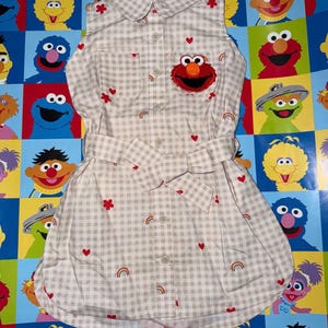 May include: A sleeveless button-down dress in a beige and white gingham check pattern. The dress features an embroidered Elmo face on the left chest pocket, along with small red heart, red flower, and rainbow motifs scattered across the fabric.