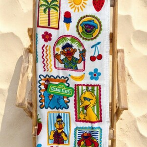May include: A white beach towel featuring a colorful Sesame Street print with characters including Ernie, Bert, Big Bird, Elmo, and Oscar the Grouch. The design includes summer icons like palm trees, suns, strawberries, ice cream, and a Sesame Street sign.
