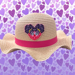 May include: A straw sun hat with a pink band and a cartoon character patch. The hat features a wide brim and a woven texture. The patch depicts a character with purple hair and a red nose. The background is a pattern of purple hearts.