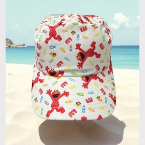 May include: A white baseball cap featuring an all-over print of the red character Elmo, small orange fish, and colorful baby bottles in blue, yellow, and purple, interspersed with red letter E characters across the crown and the curved brim.