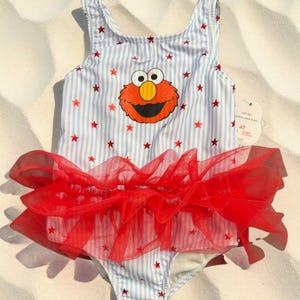 May include: A one-piece swimsuit featuring a light blue and white vertical striped pattern with small red stars throughout. The front displays a graphic of Elmo, and a layered red tulle ruffle is attached at the waist. A size 4T tag is attached.