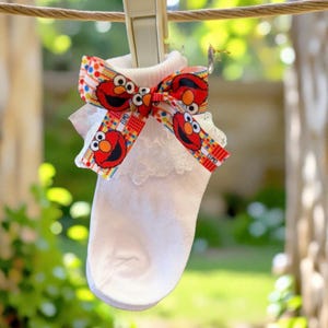 May include: A white ankle sock featuring a ruffled lace trim at the cuff and a decorative ribbon tied in a bow. The ribbon is patterned with colorful geometric shapes and multiple images of the red character Elmo from Sesame Street.