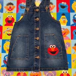 May include: A blue denim overall dress with adjustable straps and a button-down front. Features two front pockets and an embroidered Elmo patch. The dress is designed for children.