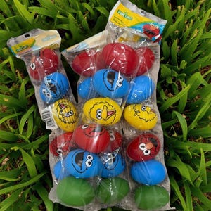 May include: A clear plastic bag filled with colorful Sesame Street character-themed Easter eggs. The eggs are red, blue, yellow, and green, featuring characters like Elmo, Cookie Monster, and Big Bird. The packaging includes the Sesame Street logo.