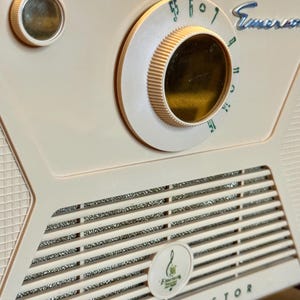 May include: Close-up of a vintage Emerson transistor radio. The radio is a light cream color with a silver speaker grill and a large tuning dial. The word "Emerson" is visible on the radio.