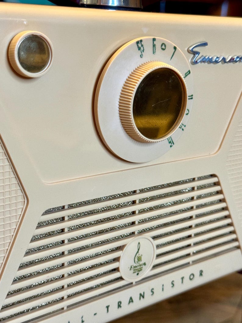 May include: Close-up of a vintage Emerson transistor radio. The radio is a light cream color with a silver speaker grill and a large tuning dial. The word "Emerson" is visible on the radio.