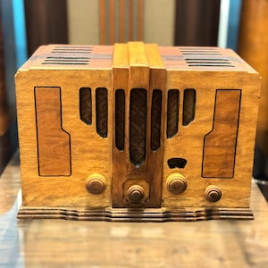 May include: A rectangular wooden radio with a light brown finish, featuring a central speaker grille with vertical slats, four circular control knobs on the front panel, and a stepped base design. The top surface includes dark brown ventilation slots.