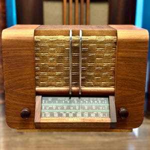 May include: A wooden radio with a textured gold fabric speaker grille, two vertical silver metal bars, and a green glass tuning dial featuring frequency scales labelled A, B, and C. Two dark brown circular knobs are positioned on the lower front.