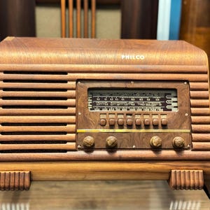 May include: A Philco tabletop radio with a horizontal wooden cabinet featuring a slatted speaker grille on the left and a central tuning dial with six push buttons. Four circular knobs are positioned below the dial, labelled bass, volume, tone, and tuning.