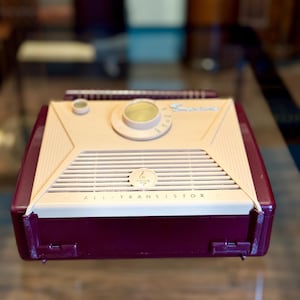 May include: A vintage, burgundy and cream-colored transistor radio. The radio has a speaker grill, a tuning dial, and the words "ALL TRANSISTOR" printed on the front. The radio has a retractable antenna.