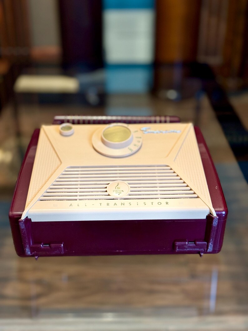 May include: A vintage, burgundy and cream-colored transistor radio. The radio has a speaker grill, a tuning dial, and the words "ALL TRANSISTOR" printed on the front. The radio has a retractable antenna.