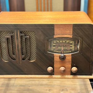 Op de afbeelding: Een rechthoekige houten radio met een tweekleurige afwerking van donkerbruine en lichtbruine houten panelen. De voorzijde bevat een luidsprekerrooster met een geometrische uitsparing, een centrale afstemknop met de tekst Stewart Warner Grenadier.