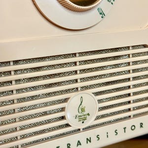 May include: Close-up of a vintage, pale pink Emerson transistor radio. The radio features a dial, speaker grill, and the words "ALL - TRANSISTOR" in green. A circular emblem with the Emerson logo is visible.