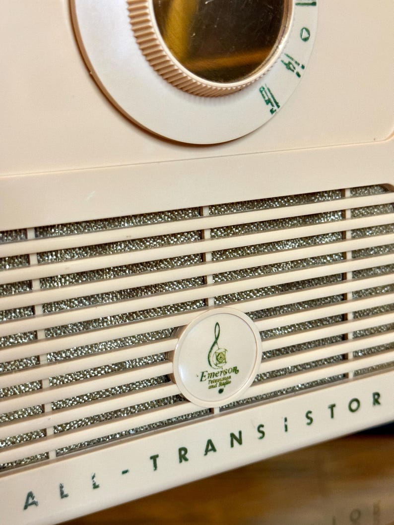 May include: Close-up of a vintage, pale pink Emerson transistor radio. The radio features a dial, speaker grill, and the words "ALL - TRANSISTOR" in green. A circular emblem with the Emerson logo is visible.
