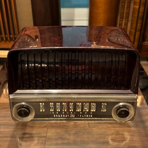 May include: A vintage General Electric radio with a dark brown, glossy finish. The radio has a black speaker grill and a dial with frequency markings. The text "GENERAL ELECTRIC" is printed on the dial. Two knobs are visible on the front.