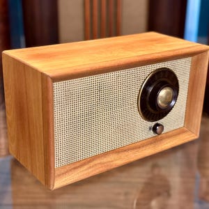 Może przedstawiać: Prostokątne radio z wykończeniem w kolorze jasnobrązowych słojów drewna oraz kremową, perforowaną metalową maskownicą głośnika. Z przodu znajduje się duże okrągłe czarne pokrętło ze złotym środkiem oraz mniejsze czarne pokrętło sterujące.