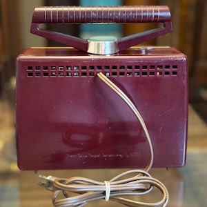 May include: A vintage burgundy-colored radio with a handle and a power cord. The radio has a rectangular shape with a series of small rectangular vents across the top. The handle is burgundy with gold-colored accents. The power cord is brown.