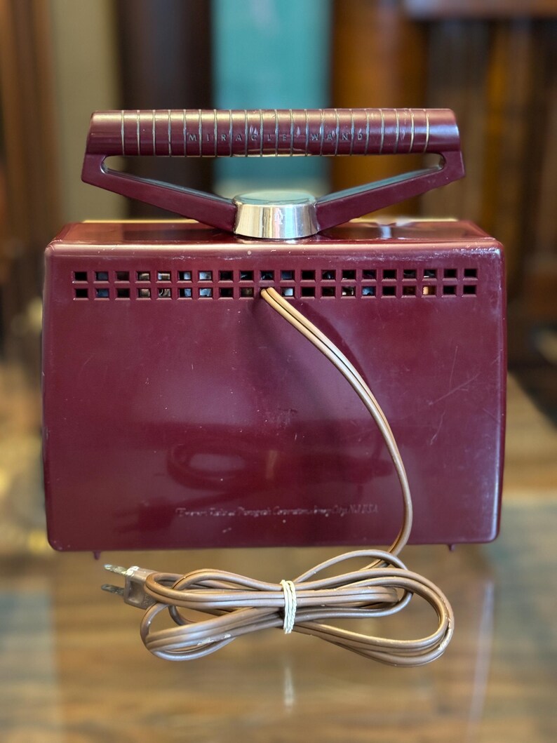 May include: A vintage burgundy-colored radio with a handle and a power cord. The radio has a rectangular shape with a series of small rectangular vents across the top. The handle is burgundy with gold-colored accents. The power cord is brown.