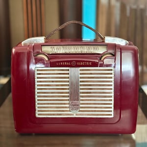 May include: A vintage General Electric Super 650 radio in a deep red casing. The radio has a brown leather handle, a dial with frequency numbers, and a speaker grille with vertical lines. The General Electric logo is visible.