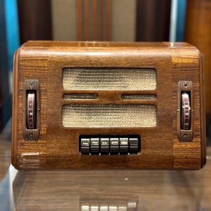 Op de afbeelding: Een vintage houten radio met een gepolijste bruine afwerking, voorzien van een centraal luidsprekerrooster van beige stof, twee verticale zijknoppen met cijfermarkeringen en een rij van vijf zwarte rechthoekige drukknoppen onder de luidspreker.