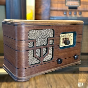 May include: A vintage wooden radio with a horizontal rectangular body and rounded edges. It features a central speaker grille covered in a textured beige fabric, a rectangular tuning dial with a cream background, and three black circular control knobs.