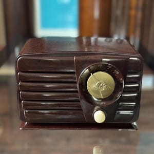 May include: A classic brown radio with a gold dial and a cream-coloured knob. The radio has a rectangular shape with horizontal lines on the front and the brand name "PHILIPS" is visible. A vintage piece of electronics.
