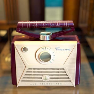 May include: Vintage Emerson radio in cream and burgundy. The radio features a dial, speaker grill, and a handle with the text "MIRACLE WAND". The radio is labeled "ALL-TRANSISTOR".