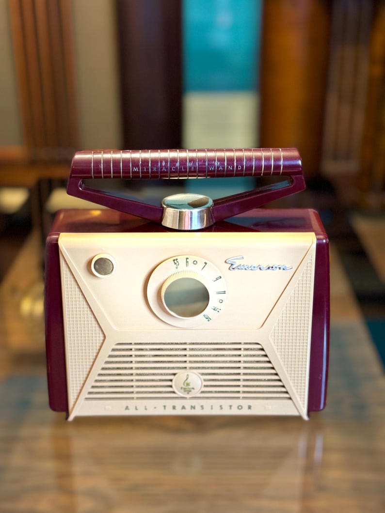 May include: Vintage Emerson radio in cream and burgundy. The radio features a dial, speaker grill, and a handle with the text "MIRACLE WAND". The radio is labeled "ALL-TRANSISTOR".